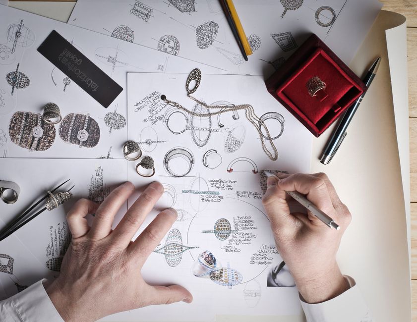 Hands sketching jewelry designs with tools and prototypes on a desk