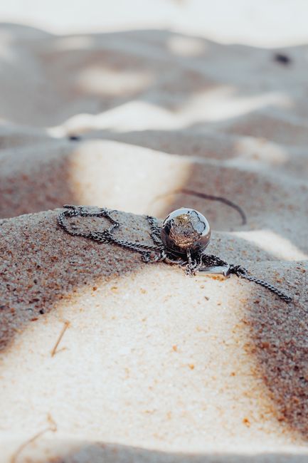 A silver necklace with a pendant lies on a sandy beach
